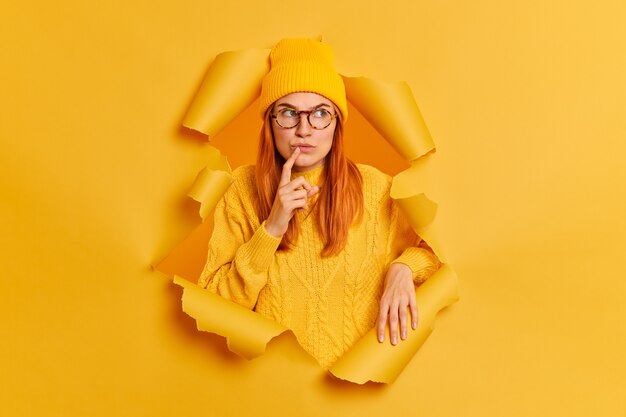 Photo of serious thoughtful woman with red hair keeps finger near lips looks pensively away recalls something or considers future plans wears hat jumper breaks through yellow paper hole. Let me think.