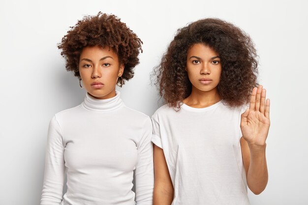 Photo of serious two Afro women have bushy curly hair, one female makes stop gesture