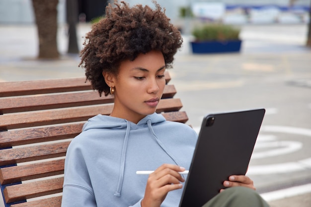 Photo of serious woman concentrated at tablet screen draws pictures with stylus poses on wooden bench dressed in hoodie spends leisure time outdoors against blurred background works freelance