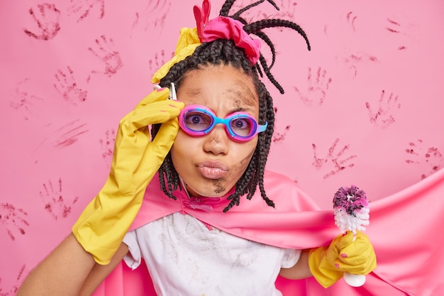 Free photo photo of serious woman housekeeper superhero cleans house holds dirty toilet brush wears goggles pink cape and rubber gloves pretends having superpower busy doing housework or household chores