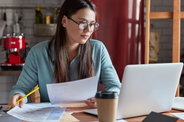 Photo of serious young European woman reads information from paper documents, checks on modern laptop computer, poses against kitchen interior,