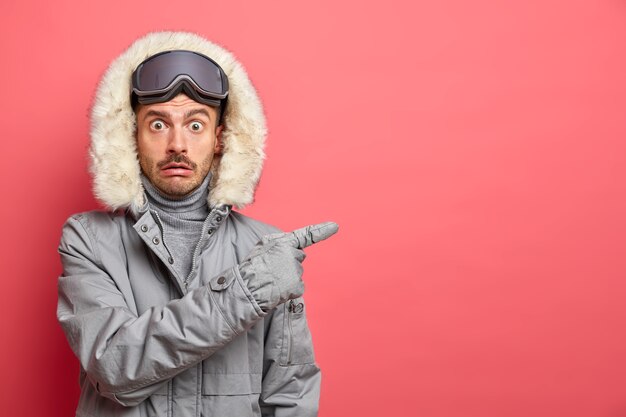 겨울 옷을 입은 충격을받은 감정적 인 유럽 남자의 사진은 스키 고글을 착용하고 빈 공간을 가리켜 오른쪽에 방향을 제공합니다.