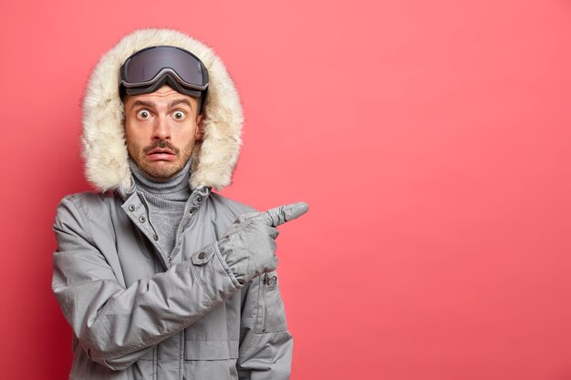 Photo of shocked emotional European man dressed in winter clothes wears ski goggles and points away on blank space gives direction on right.