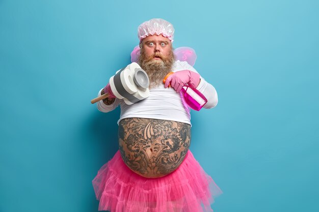 Photo of shocked plump bearded man pretends being fairy of purity cleans something holds bottle of detergent and plunger has big tattooed belly