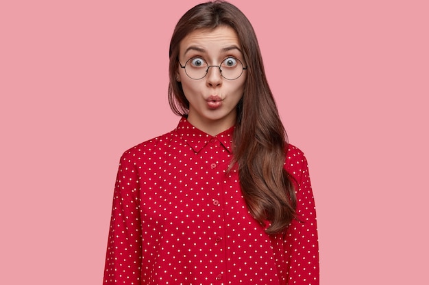 Photo of shocked woman in stupor, keeps lips round, has distrubing situation, dressed in fashionable polka dot blouse, wears round spectacles