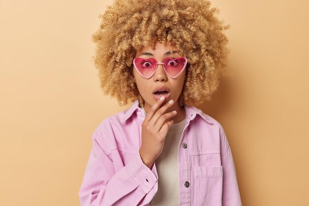 Photo of shocked woman with curly hair gasps from wonder keeps jaw dropped wears heart shaped sunglasses pink jacket cannot believe in amazing news isolated over beige background Omg concept