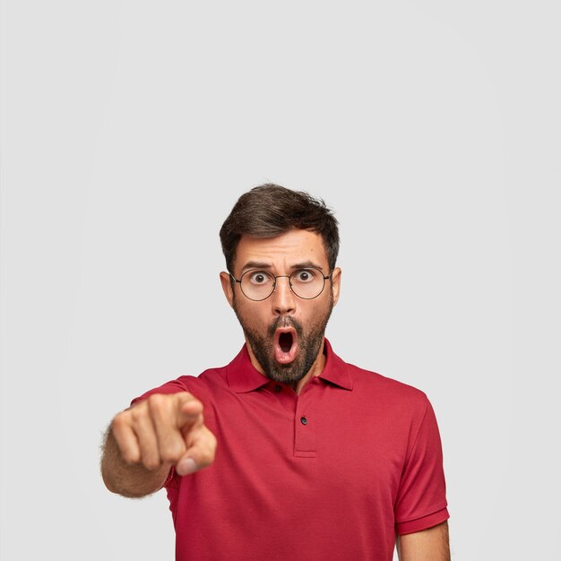 Photo of shocked young male with bristle, has stunned expression, points directly with index finger, wears casual red t-shirt, stands against white wall, notices something awful in front
