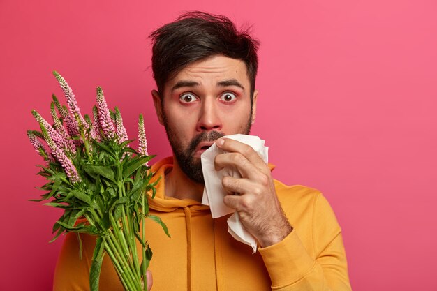 Фотография шокированного молодого человека с аллергией на весенние цветы или растения, астматическим заболеванием, покраснением вокруг носа, держащим платок, изолированным на розовой стене. Здравоохранение, сенная лихорадка, концепция болезни