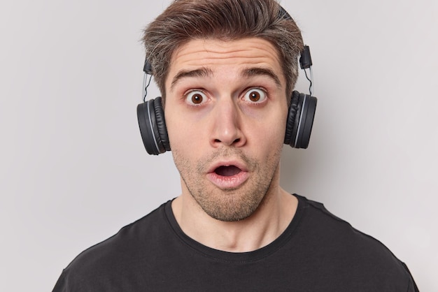 Photo of shocked young man listens radio online via sereo headphones keeps mouth opened feels stunned dressed in casual black t shirt reacts on something stunning isolated over white background