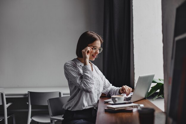 Photo of short-haired business woman in glasses and white blouse sitting in workplace and working in laptop.