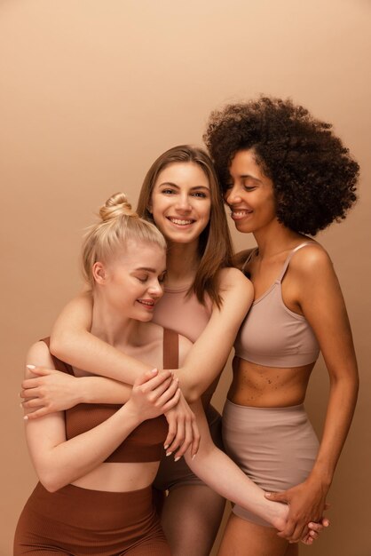 写真は、ベージュの背景で一緒に時間を過ごしているトップとレギンスを着ている3人の若い多様な女の子を示しています