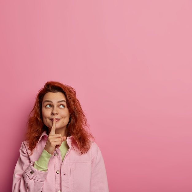 Free photo photo of silent woman makes silence gesture, keeps index finger over lips, looks above, gossips about something