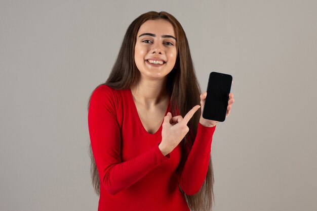 Photo of smiling adorable girl in red sweatshirt holding cellphone on gray wall.