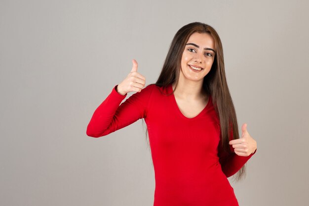 Photo of smiling adorable girl in red sweatshirt standing and giving thumbs up on gray wall.