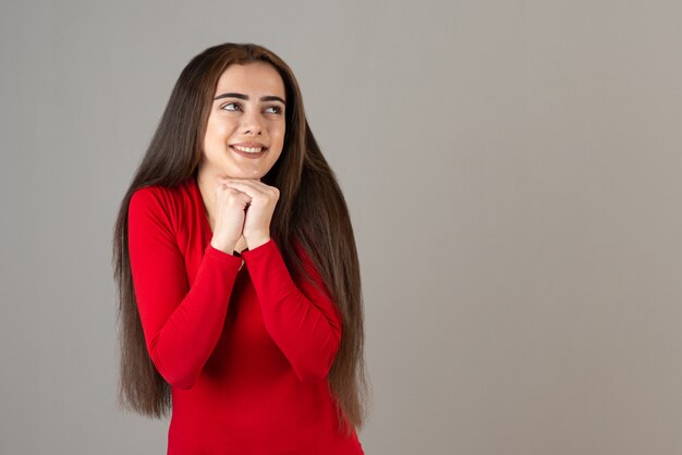 灰色の壁に立っている赤いスウェットシャツの笑顔の愛らしい女の子の写真。