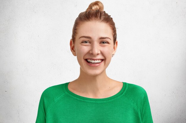 머리 매듭을 짓고 웃는 귀여운 유럽 여성의 사진은 밝은 녹색 스웨터를 입고 넓은 미소와 하얀 이빨을 가지고 있으며 연인과 잊을 수없는 하루를 보내고 기뻐합니다.