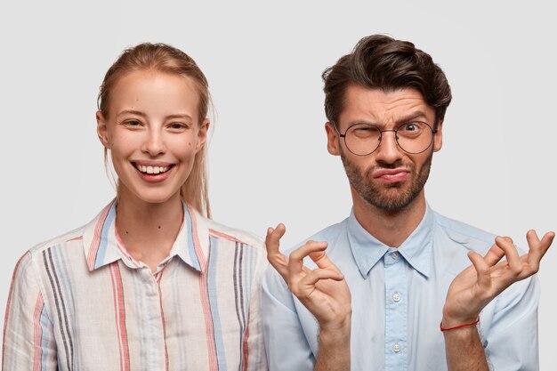 Photo of smiling light haired woman, expresses happiness, discontent unshaven man with gloomy expression, raises eyebrows in dislike, spreads hands, isolated over white wall. Emotions, reaction