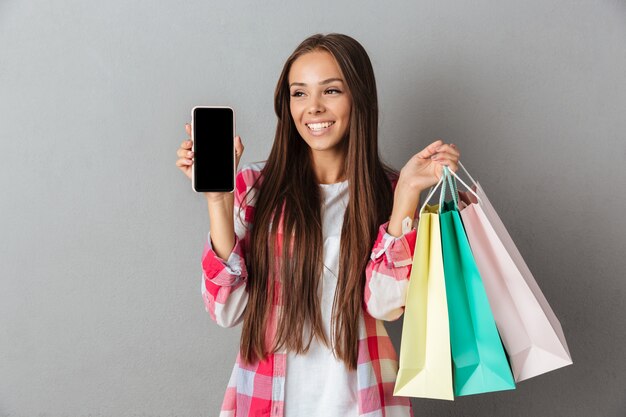 Фотография улыбающейся молодой брюнетки, держащей хозяйственные сумки, показывающей пустой экран мобильного телефона и отводящей взгляд