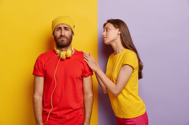 Photo of sorrowful unshaven man has trouble, wears yellow hat and red t shirt, smirks face with displeasure, caring girlfriend touches his shoulder, tries to calm and help in difficult situation