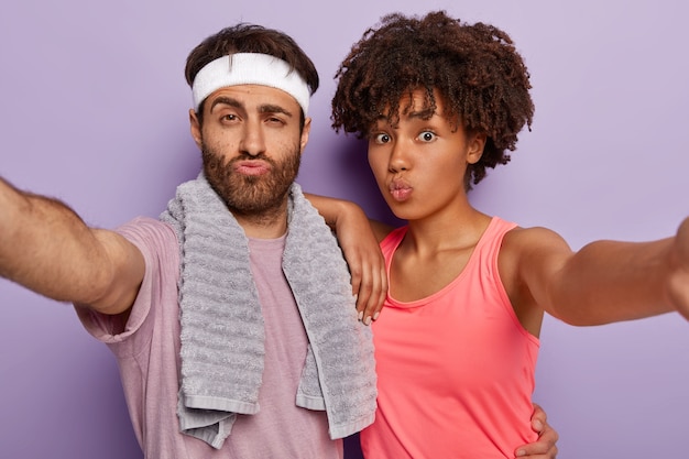 На фото спортивная женщина и мужчина протягивают руки, делают селфи-портрет, держат губы сложенными, одет в активную одежду, мягкое полотенце на плечах.