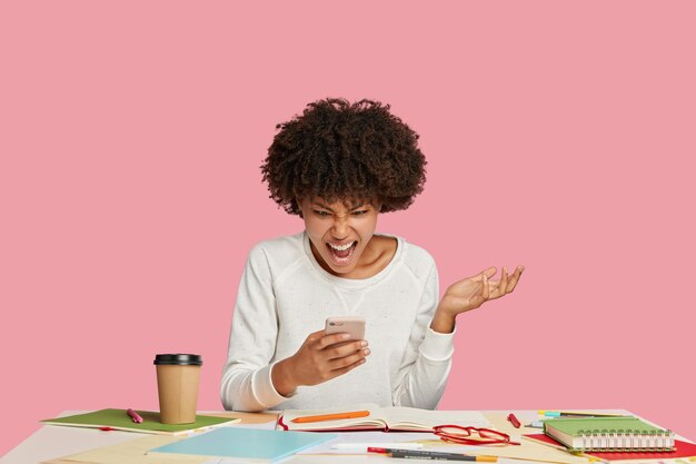 Photo of stressful black female worker or secretary looks at screen of smart phone with irritation