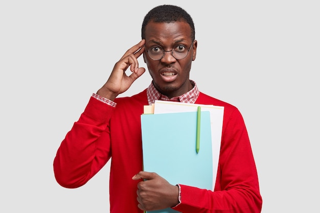 Photo of stressful displeased dark skinned young guy kepes hand on temple, frowns face, dressed in red elegant sweater