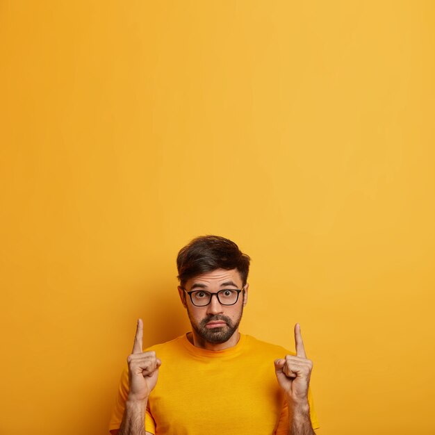 Photo of stunned man shows product, recommends service with amazed expression, stares with bugged eyes, indicates on blank space upwards, wears optical glasses, isolated on yellow wall