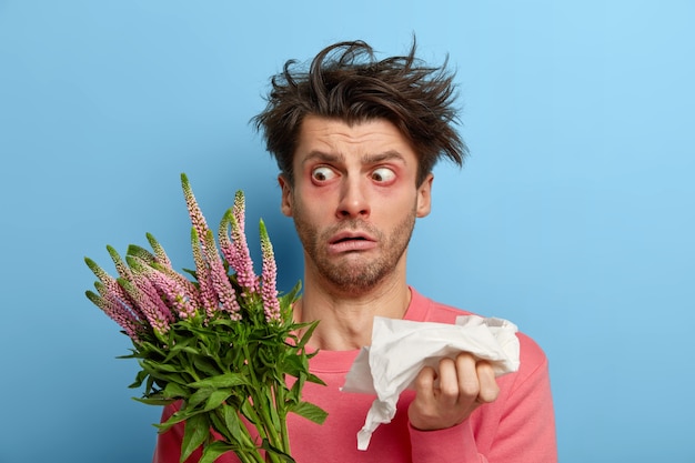 Photo of stunned man stares at plant with widely opened eyes, has messy hair, holds napkin as sneezes all time, allergic reaction to pollen and flowering, feels sick and exhausted of being ill