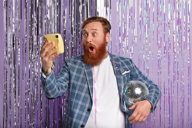 Free photo photo of stupefied caucasian man with red hair and beard, holds disco ball under arm, looks surprisingly, makes photo of himself, wears stylish attire, has fun on office party