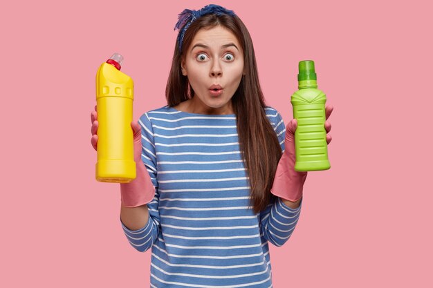 Photo of stupefied Caucasian woman from cleaning service, carries necessary supplies, wears rubber gloves, dressed in striped sweater