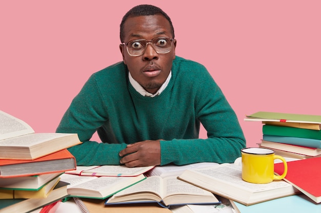 Фотография ошеломленного темнокожего мужчины, смотрящего широко открытыми глазами, одетого в зеленый свитер, окруженного большим количеством литературы, пишет курсовую работу.