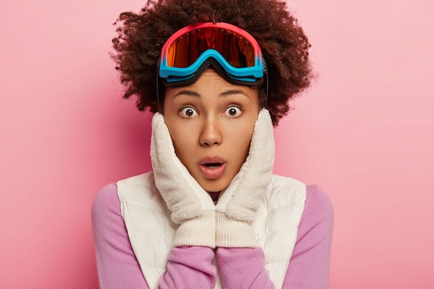 Free photo photo of stupefied emotional curly female looks with shocked face expression at camera, keeps both hands on cheeks, wears ski glasses for snowboarding, isolated over rosy studio wall