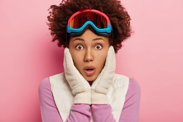 Фотография ошеломленной эмоциональной кудрявой женщины с шокированным выражением лица в камеру, держит обе руки на щеках, носит лыжные очки для сноуборда, изолирована на розовой стене студии
