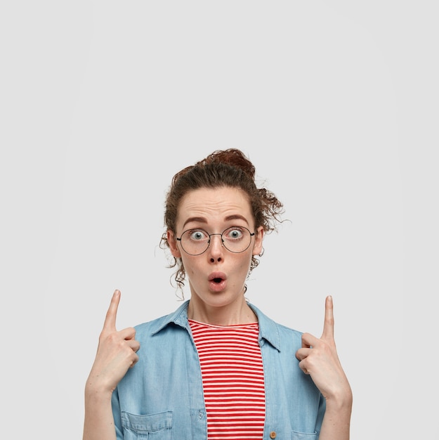 Photo of stupefied emotive freckled woman with dark hair, wears round spectacles