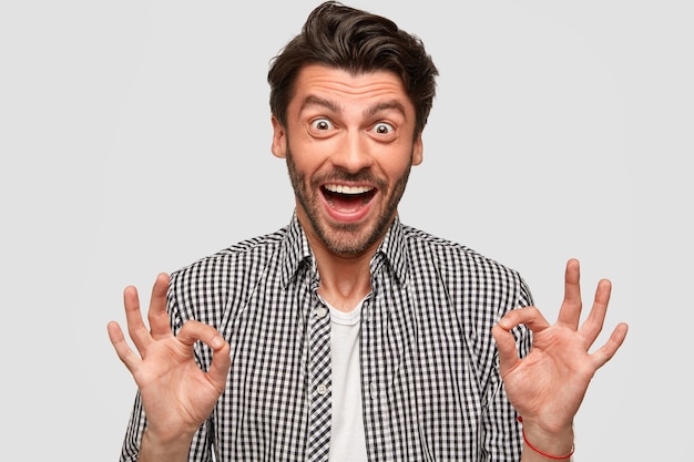 Photo of stylish bearded young male has trendy haircut, makes okay gesture with both hands, dressed in checkered shirt, stares with eyes, isolated over white wall. Body language concept