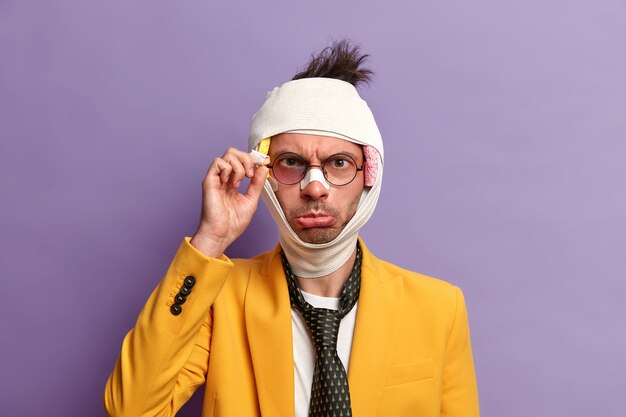 Photo of sullen displeased man with bruise near eye, hematoma and brain concussion, wears bandage, formal suit and tie, beaten by cruel people, isolated on purple wall. Health problem concept