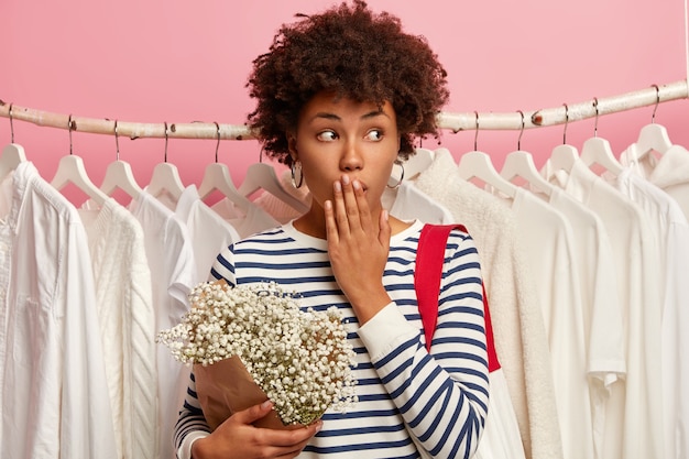 Фотография удивленной покупательницы афроамериканского происхождения прикрывает рот и смотрит в сторону, одетая в полосатую одежду, держит букет, стоит на фоне белых нарядов, висящих в один ряд на рельсах, изолирована на розовом