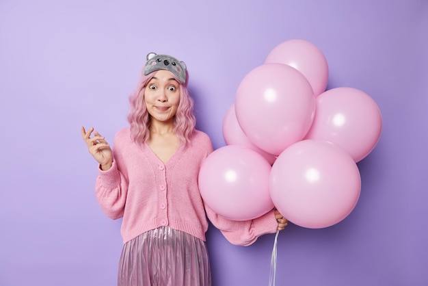 Photo of surprised Asian pink haired woman looks wondered prepares for holiday wears festive clothes holds bunch of inflated helium balloons isolated over purple background Party time concept