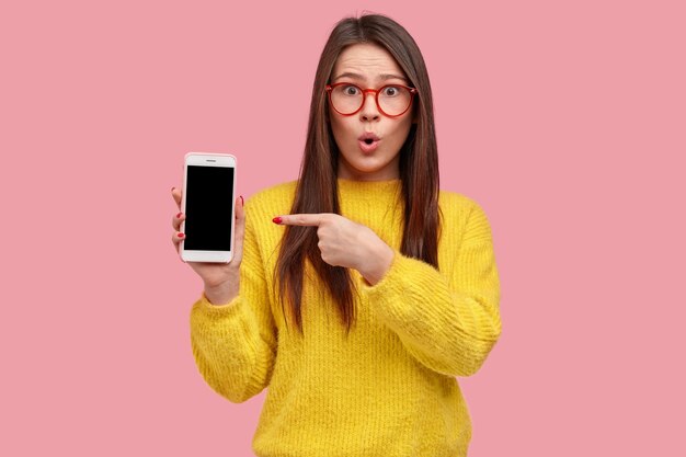 Photo of surprised attractive woman points at blank empty screen of modern gadget, keeps jaw dropped, wears yellow clothes