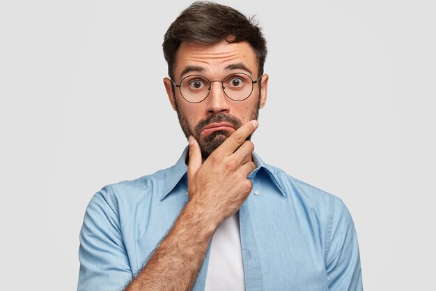 Photo of surprised bearded male holds chin and looks in hesitation, wonders latest news, wears spectacles and elegant blue shirt, isolated over white wall. People and facial expressions concept