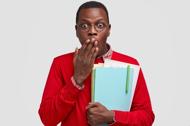 Photo of surprised black man covers mouth with palm, has scared expression, carries textbooks, dressed in red jumper and spectacles