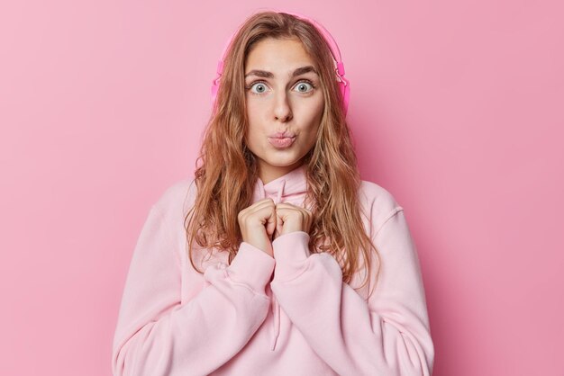 Photo of surprised blue eyed woman keeps hands together has lips rounded long dark hair dressed in casual sweatshirt listens music via headphones isolated over pink background Hobby concept
