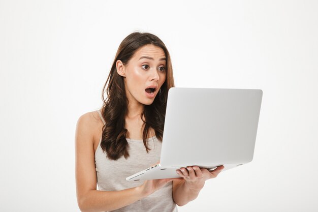 Photo of surprised brunette woman looking on screen of new notebook being excited or thrilled, isolated over white wall