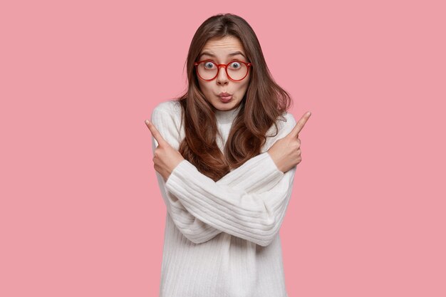 Photo of surprised Caucasian lady crosses hands over chest, points with both index fingers, hesitates between two objects