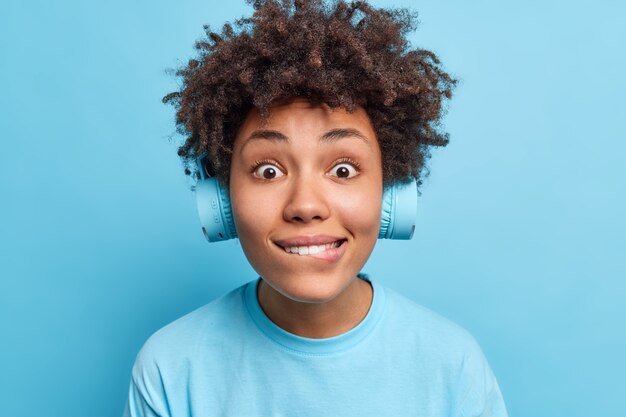 Photo of surprised curious Afro American woman bites lips looks directly  wears wireless headphones dressed in casual t shirt isolated over blue wall. People leisure lifestyle