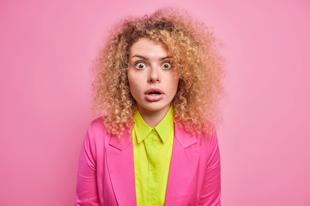 Photo of surprised curly haired young European woman cannot believe in shocking news holds breath from amazement dressed in formal bright clothes isolated over pink wall. Emotions concept