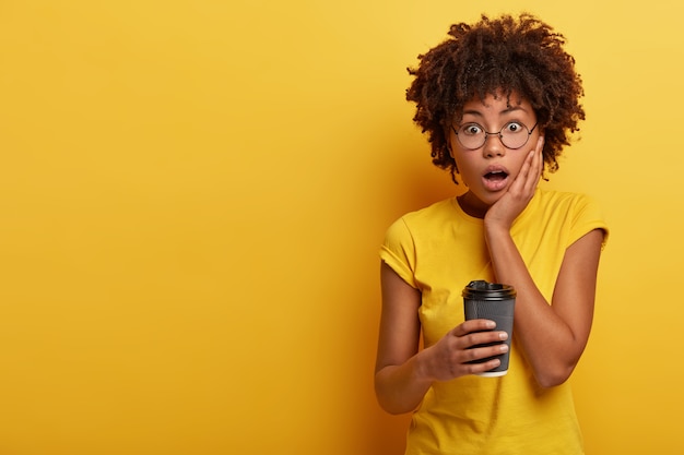 Photo of surprised dark skinned woman looks in stupor at camera, keeps hand on cheek