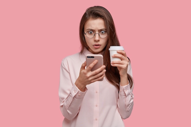 Photo of surprised dark skinned woman looks with stunned expression at screen of cell phone, has problem, recieved unpleasant news