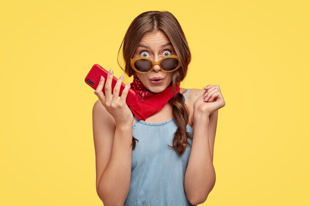 Photo of surprised emotive dark haired woman in trendy shades holds cell phone, hears something astonishing, wears red bandana, models over yellow wall. People, reaction and style concept.