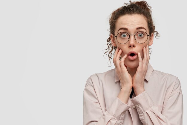 Photo of surprised European female gasps and gasps breath in shock, opens eyes widely, wears casual shirt, stands against white wall